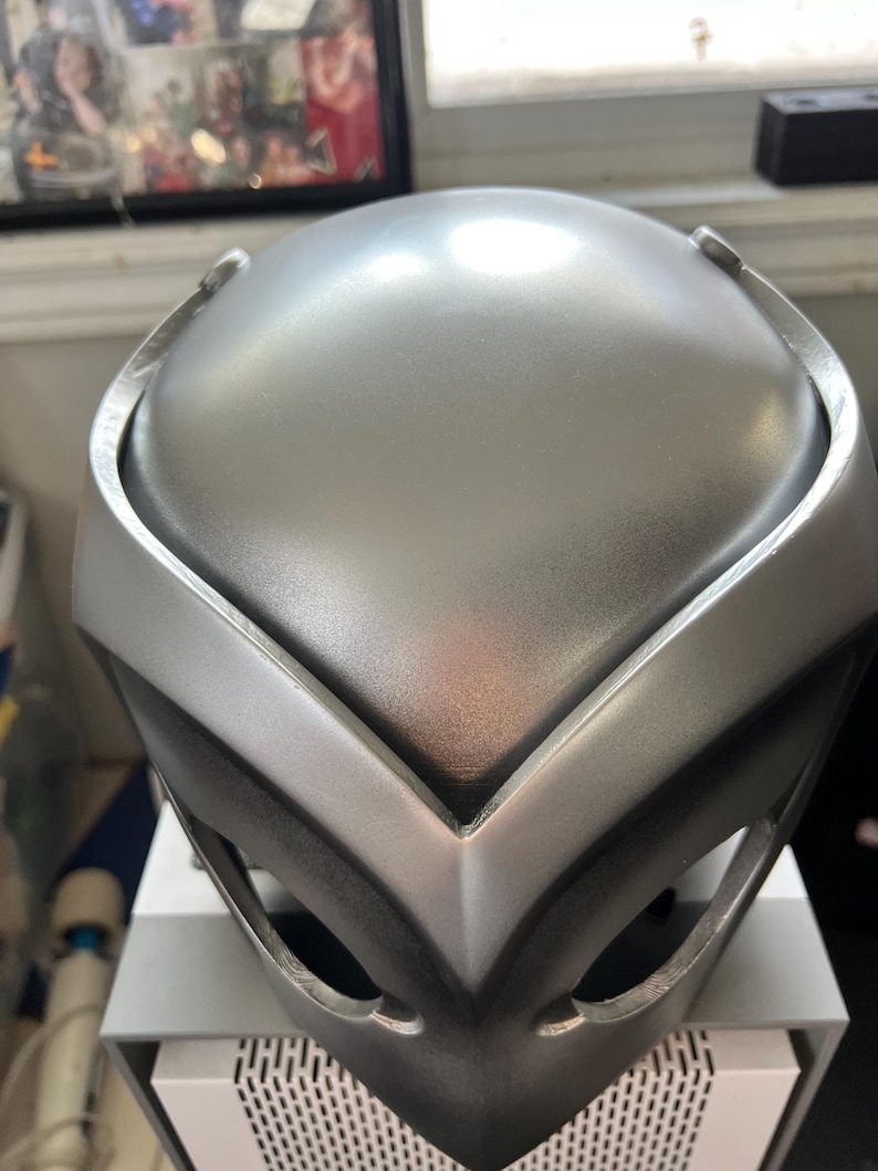 May include: A silver-colored helmet with a V-shaped design on the front. The helmet has a smooth, metallic finish.