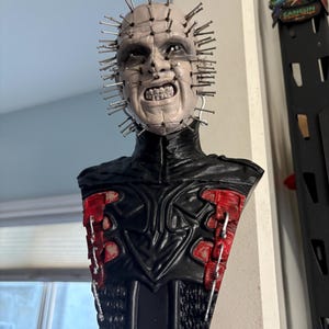 May include: A detailed bust of a character with a pale face, numerous metal pins, and a grimacing expression. The figure wears a black outfit with red accents, showcasing a horror theme.