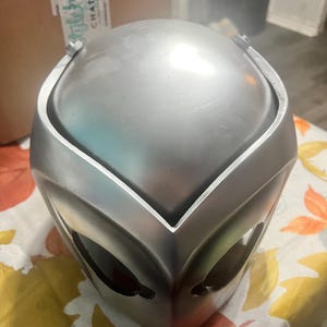 May include: A silver helmet with a smooth, shiny finish. The helmet has a pointed, V-shaped design on the front and two eye holes.