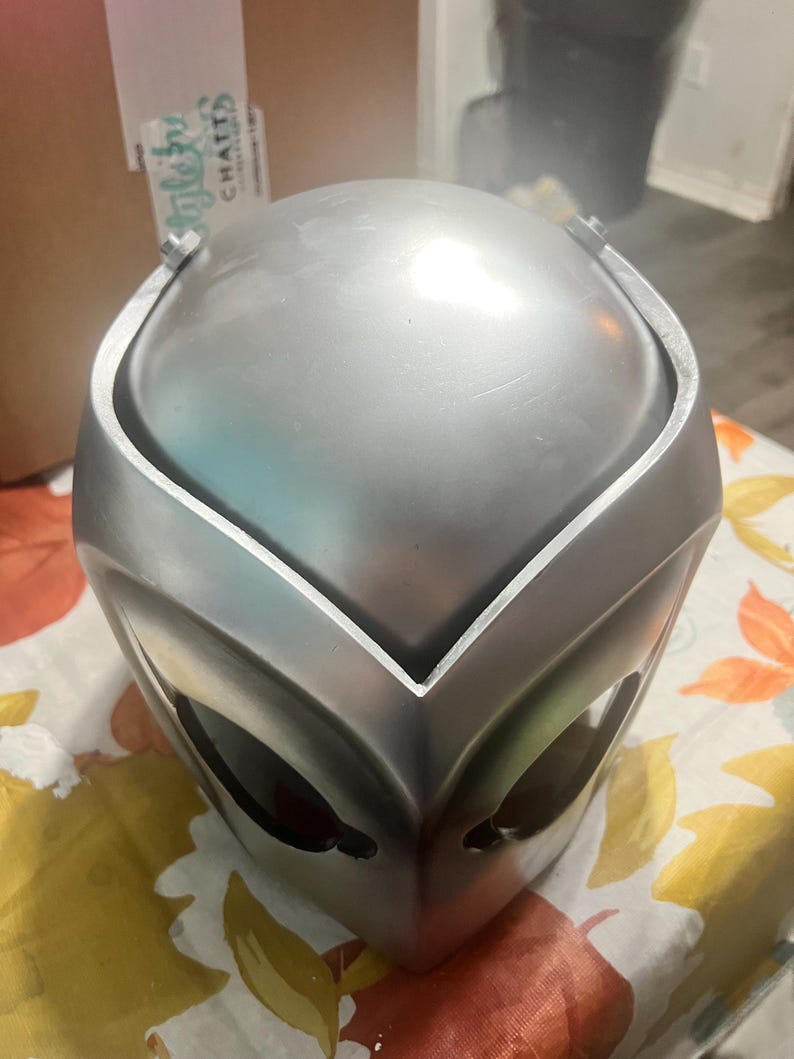 May include: A silver helmet with a smooth, shiny finish. The helmet has a pointed, V-shaped design on the front and two eye holes.