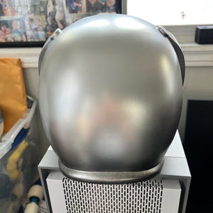 May include: A silver helmet with a smooth, shiny finish. The helmet has a rounded shape and a slightly raised rim around the bottom edge.