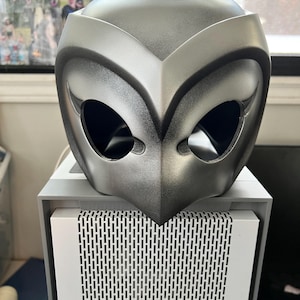 May include: A silver-coloured plastic mask with a bird-like design. The mask has two large eye holes and a pointed beak.