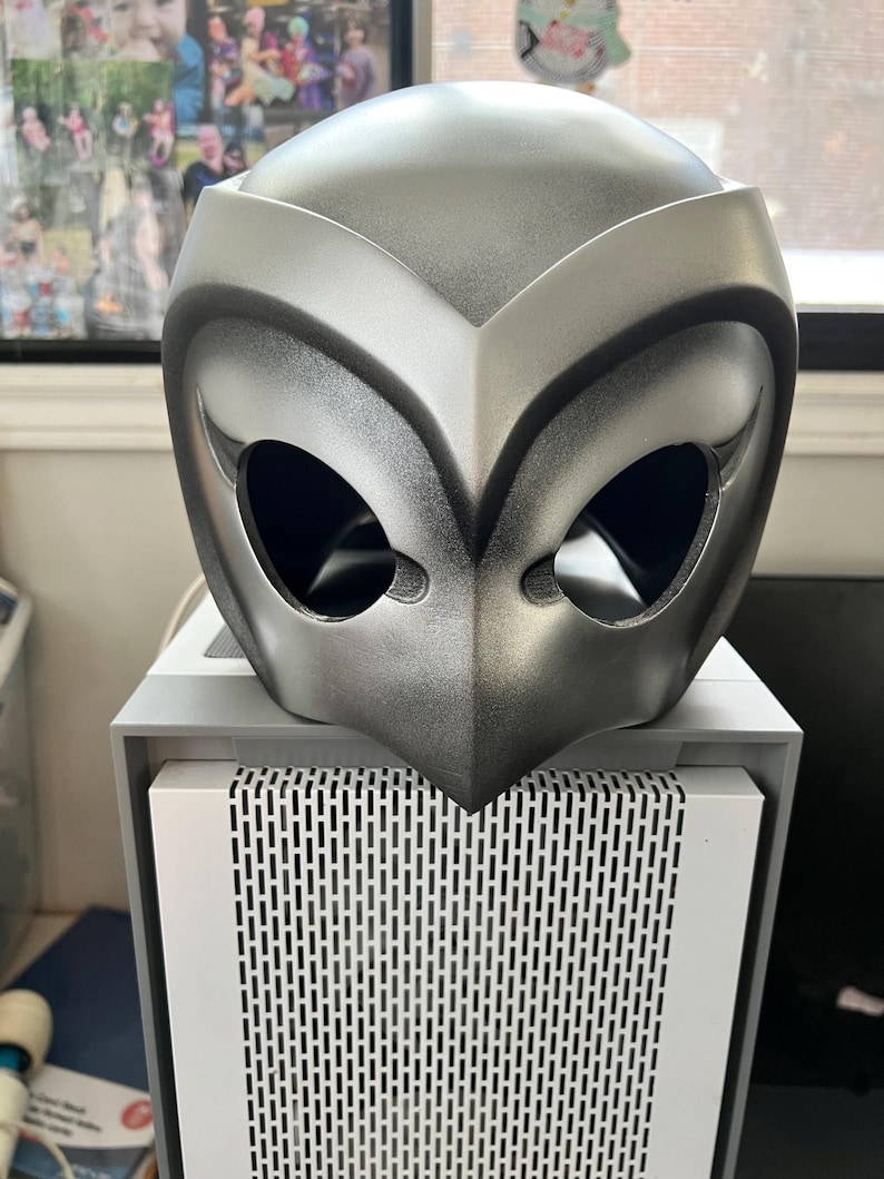 May include: A silver-colored plastic mask with a bird-like design. The mask has two large eye holes and a pointed beak.