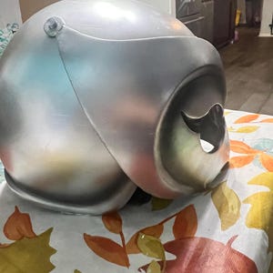 May include: A silver helmet with a visor and a hole on the side. The helmet is sitting on a table with a floral pattern.