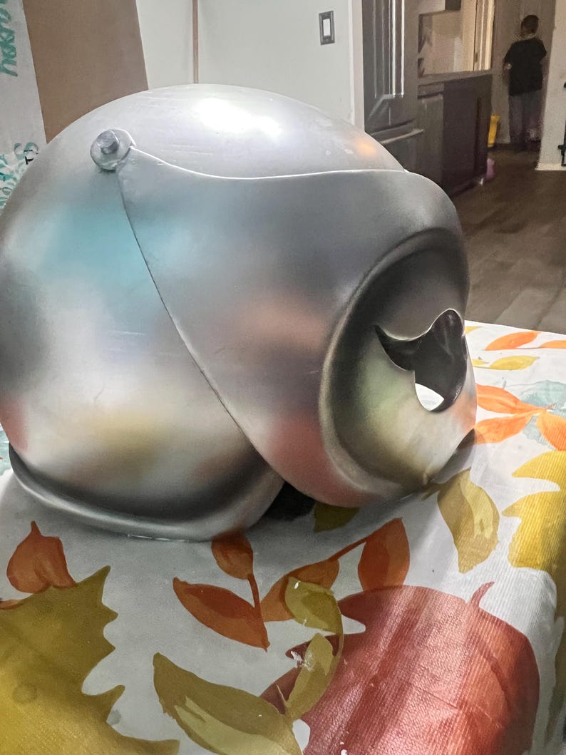 May include: A silver helmet with a visor and a hole on the side. The helmet is sitting on a table with a floral pattern.