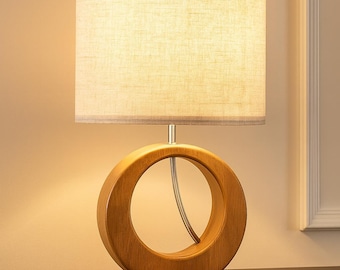 May include: A wooden table lamp with a beige fabric shade. The lamp base features a circular design with a light brown finish. The lamp is placed on a dark brown wooden surface, creating a warm and inviting ambiance.