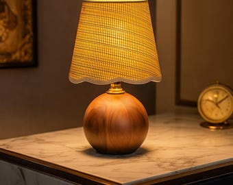 May include: A small, decorative table lamp with a wooden base and a textured, yellow lampshade. The lamp has a round, spherical base and a scalloped edge on the lampshade. A gold-colored clock is visible in the background.