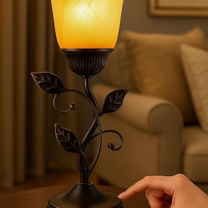 May include: A decorative table lamp with a floral design. The lamp features a warm yellow glass shade and a dark bronze-colored stem with leaf accents. The lamp is turned on with a touch of a finger.