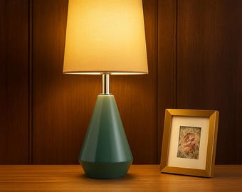 May include: A teal table lamp with a beige lampshade and a gold-colored metal stem. A small gold picture frame with a floral image sits to the right of the lamp. The lamp is on a wooden surface.