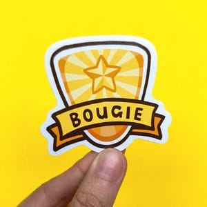 Puede incluir: Una pegatina con un diseño de escudo con una estrella y líneas radiantes en tonos amarillos y naranjas. Una pancarta en la parte inferior dice "BOUGIE" en letras negras en negrita. La pegatina se sostiene sobre un fondo amarillo brillante.