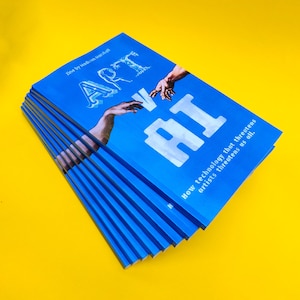 May include: A stack of blue zines with the title "ART vs AI" and the text "How technology that threatens artists threatens us all." The cover features a painting of two hands reaching towards each other.