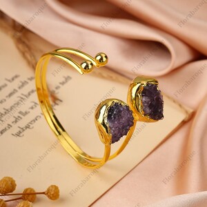 May include: A gold-coloured cuff bracelet featuring two rough-cut, purple amethyst stones. The bracelet has a curved design with a smooth, polished finish. The amethyst stones are set in gold-coloured bezels.
