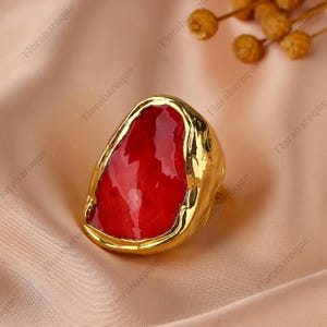 May include: A gold-toned ring with a large, irregular shape. The ring features a vibrant red, stone-like center. The ring is set against a soft, peach-colored fabric, with the brand name "Florabaroque" visible in the background.