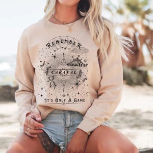 May include: Beige sweatshirt with a circular graphic design featuring the text "REMEMBER CARAVAL" and "It's ONLY A GAME". The design includes a compass, stars, and a banner. The sweatshirt is a neutral color.
