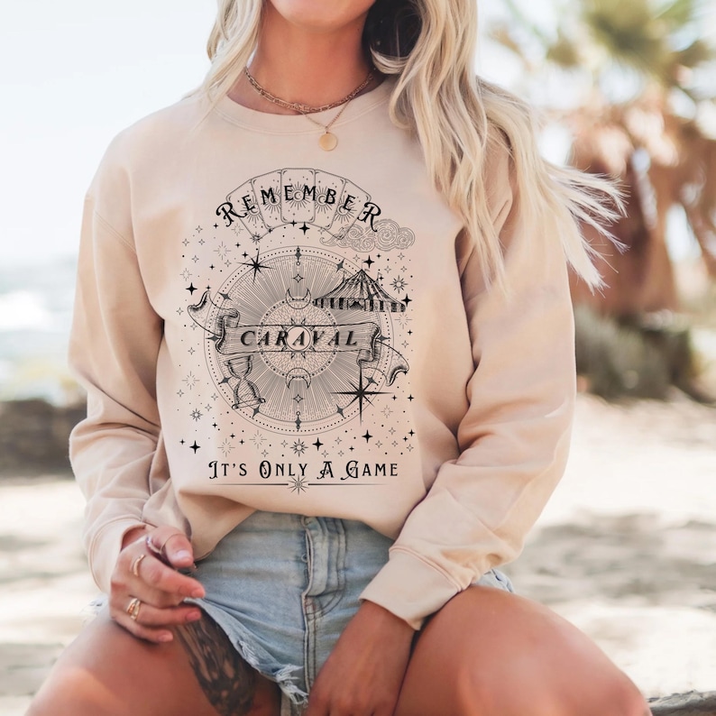 May include: Beige sweatshirt with a circular graphic design featuring the text "REMEMBER CARAVAL" and "It's ONLY A GAME". The design includes a compass, stars, and a banner. The sweatshirt is a neutral color.