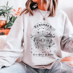 May include: A light gray sweatshirt with a black and white graphic design. The design includes the words "REMEMBER CARAVAL" on a banner, a compass, stars, and the phrase "IT'S ONLY A GAME".