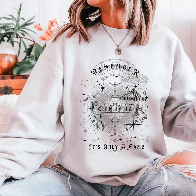May include: A light gray sweatshirt with a black and white graphic design. The design includes the words "REMEMBER CARAVAL" on a banner, a compass, stars, and the phrase "IT'S ONLY A GAME".