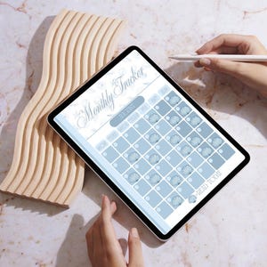 May include: A digital tablet showing a "Monthly Tracker" calendar for August, with a light blue seashell design. A wooden decorative piece and a hand holding a stylus are also visible.