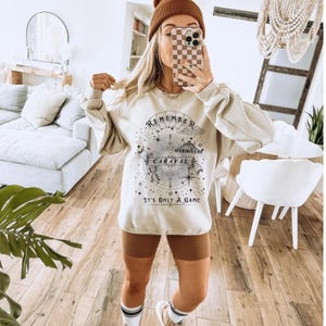 May include: Beige sweatshirt with a graphic design featuring the text "REMEMBER CARAVAL" and "IT'S ONLY A GAME". The design includes a compass rose, stars, and a circus tent. The sweatshirt has long sleeves.