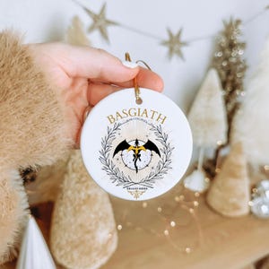 May include: A white ceramic ornament with a gold string, featuring the text "BASGIATH WAR COLLEGE" and a black and gold dragon design. The ornament is held in a hand, with holiday decorations in the background.