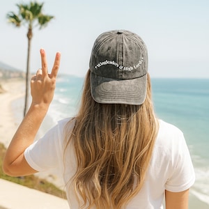 May include: A gray baseball cap with the text "Wingleaders & High Lords" in white. The cap is worn by a person with long blonde hair, standing in front of a beach and ocean. The person is wearing a white t-shirt and making a peace sign.