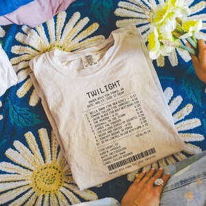 Op de afbeelding: Crèmekleurig T-shirt met het woord "TWILIGHT" bovenaan, gevolgd door een lijst met titels en tijden. Het shirt heeft ook een barcode en bestelgegevens. Het shirt is opgevouwen op een blauw bloemenpatroon.