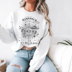 May include: White sweatshirt with a black and white graphic design. The design includes the word "Remember" above a compass rose, the word "Caraval" on a banner, and the phrase "It's Only A Game".