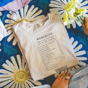Könnte beinhalten: Cremefarbenes T-Shirt mit dem Aufdruck "MANACLED" oben sowie einer Liste von Titeln und Zeiten. Das Shirt liegt auf einer blau gemusterten Decke. Das Shirt ist XL-TG/20.
