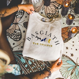 Könnte beinhalten: Cremefarbenes Sweatshirt mit dem Text "BASGIATH WAR COLLEGE DRAGON RIDERS" in Schwarz, Blau und Gold. Das Design zeigt drei Drachensilhouetten. Das Sweatshirt liegt gefaltet auf einem Stoff mit Schmetterlingsmuster.