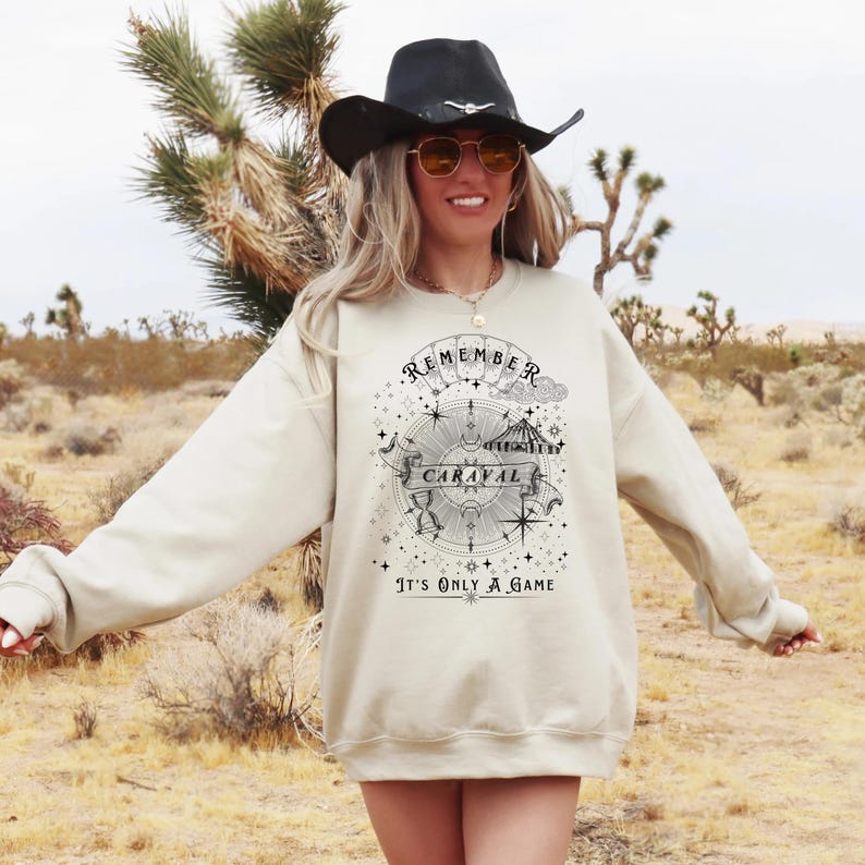 May include: A cream-colored sweatshirt with the text "REMEMBER CARAVAL" in a circular design with stars and a compass. Below the design is the text "IT'S ONLY A GAME". The sweatshirt is worn by a person wearing a black hat and sunglasses.