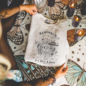 May include: A cream-colored sweatshirt with a crew neck. The front features a circular graphic with the words "REMEMBER CARAVAL" and "It's ONLY A GAME". The design includes stars, a compass, and an hourglass. The sweatshirt is folded.