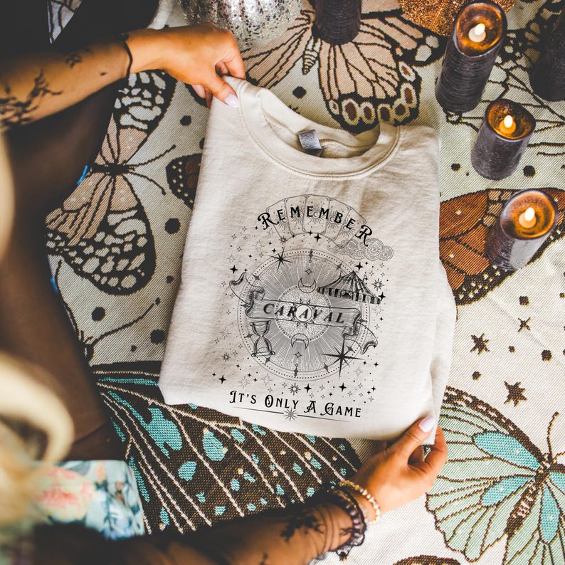 May include: A cream-colored sweatshirt with a crew neck. The front features a circular graphic with the words "REMEMBER CARAVAL" and "It's ONLY A GAME". The design includes stars, a compass, and an hourglass. The sweatshirt is folded.