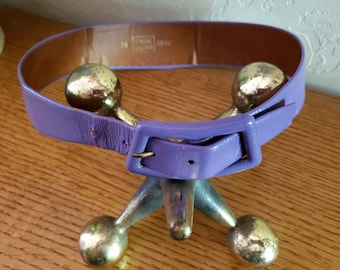 May include: A vintage lavender belt with a square buckle, displayed on a decorative metal stand. The belt is made of a shiny material and has a brown interior. The stand has three legs and gold-colored ball accents.