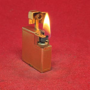 May include: A gold-coloured, rectangular lighter with a textured surface and a flame. The lighter is lit, with the flame extending upwards. The lighter has a hinged top and a small wheel for ignition. The background is a solid red.