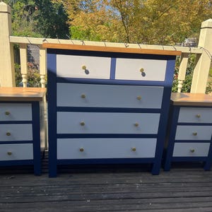 May include: A bedroom furniture set consisting of one large dresser and two matching nightstands. Each piece features a deep navy blue frame, white drawer fronts, a natural wood top surface, and small circular gold-toned metal knobs.