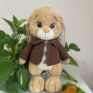 May include: A plush bunny toy with a brown jacket and large, expressive eyes. The bunny is light brown with a white chest and inner ears. A crocheted carrot with green leaves sits nearby. The toy is designed to look like a children's character.