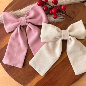 Könnte beinhalten: Zwei Stoffhaarbänder, eines rosa und eines beige, zu einer großen Schleife gebunden. Die Schleifen werden auf einer Holzoberfläche präsentiert.