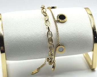 Puede incluir: Dos pulseras doradas exhibidas en un soporte cilíndrico blanco con detalles dorados. Una pulsera presenta un diseño de eslabones de cadena, mientras que la otra tiene una cadena delicada con colgantes circulares negros y blancos. Las pulseras son de metal brillante.