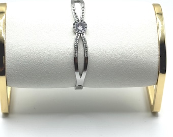 May include: A silver-colored bracelet with a central circular design, encrusted with small, clear gemstones. The bracelet has an open design with a hinge closure. The bracelet is displayed on a white surface with gold-colored supports.
