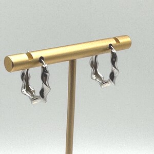 May include: Silver-coloured hoop earrings with a twisted design. The earrings are displayed on a gold-coloured earring stand against a white background. The earrings have a unique, wavy shape.