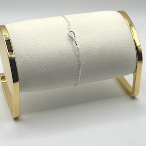 May include: A delicate silver bracelet with a knot design. The bracelet is displayed on a white, cylindrical jewellery holder with gold-coloured accents. The background is plain white.