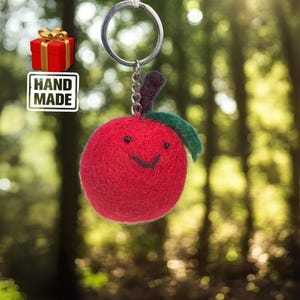 May include: A handmade red apple keyring with a smiling face, green leaf, and metal keyring. The keyring is attached to a chain and a silver ring. The image also includes a red gift box with a gold bow and a "HAND MADE" label.