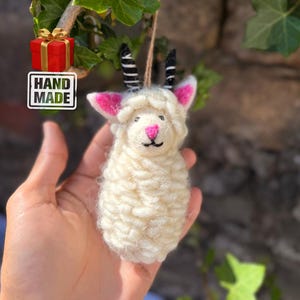 May include: Handmade, cream-coloured sheep ornament with pink ears and nose, black horns, and a black embroidered mouth. The ornament is suspended from a twine string. A red gift box and a "HAND MADE" sticker are in the background.