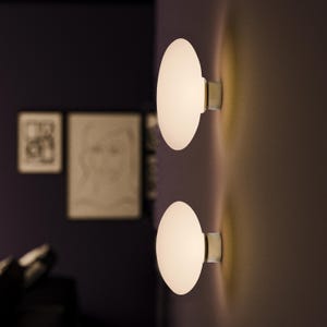 Könnte beinhalten: Zwei weiße, runde Wandleuchten mit verchromter Oberfläche. Die Leuchten sind eingeschaltet und strahlen ein warmes Licht ab.
