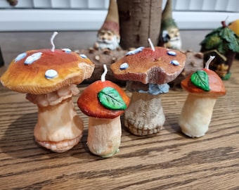 May include: Four mushroom-shaped candles with white wicks. The candles are decorated with red and brown paint and green leaves. The candles are arranged on a wooden surface.