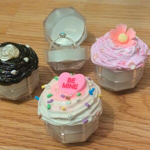 Könnte beinhalten: Drei cupcakeförmige Ringboxen. Eine ist schwarz mit einer weißen Rose oben, eine ist rosa mit einer weißen Blume oben und eine ist weiß mit einem rosa Herzen, auf dem "Be Mine" steht.