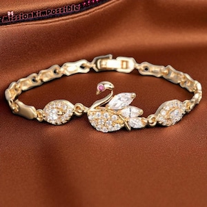 Puede incluir: Pulsera dorada con diseño de cisne. El cisne está adornado con cristales transparentes y brillantes, con una pequeña piedra roja para el ojo. La pulsera tiene un diseño de cadena delicado con detalles de cristal, ideal para añadir elegancia.