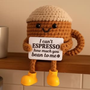 May include: A crocheted coffee cup character with a tan top and brown body, black eyes, and a smiling face. It holds a sign that reads "I can't ESPRESSO how much you bean to me." The character wears yellow boots.