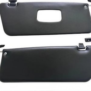 May include: Two dark grey car sun visors. One visor has a rectangular cutout. Both visors have a mounting bracket and a small, rectangular clip. The visors are made of a smooth, matte material.