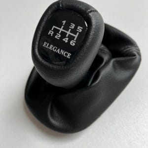 May include: Black leather gear shift knob with a white gear pattern and the word "ELEGANCE" printed on the front. The gear pattern includes numbers and letters for the gears. The knob is designed for a manual transmission.